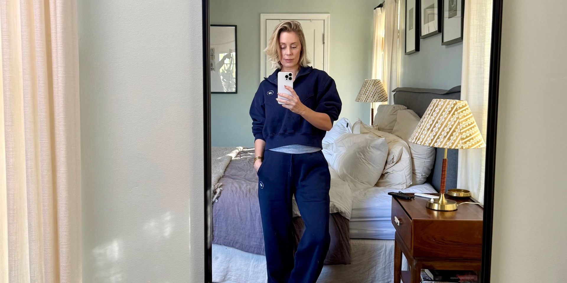 Person in a navy tracksuit taking a mirror selfie in a cozy bedroom with a neatly made bed and a nightstand.