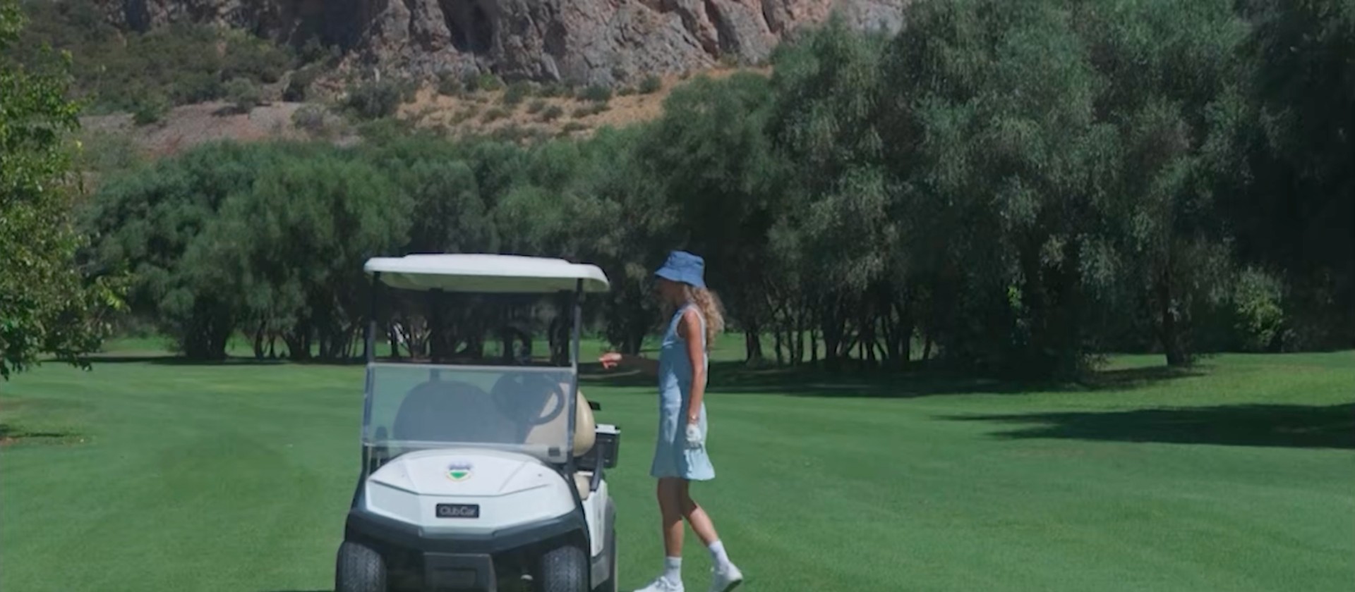 Golfer in blue outfit standing by a golf cart on a lush green course, with a rocky mountain backdrop under a clear sky.