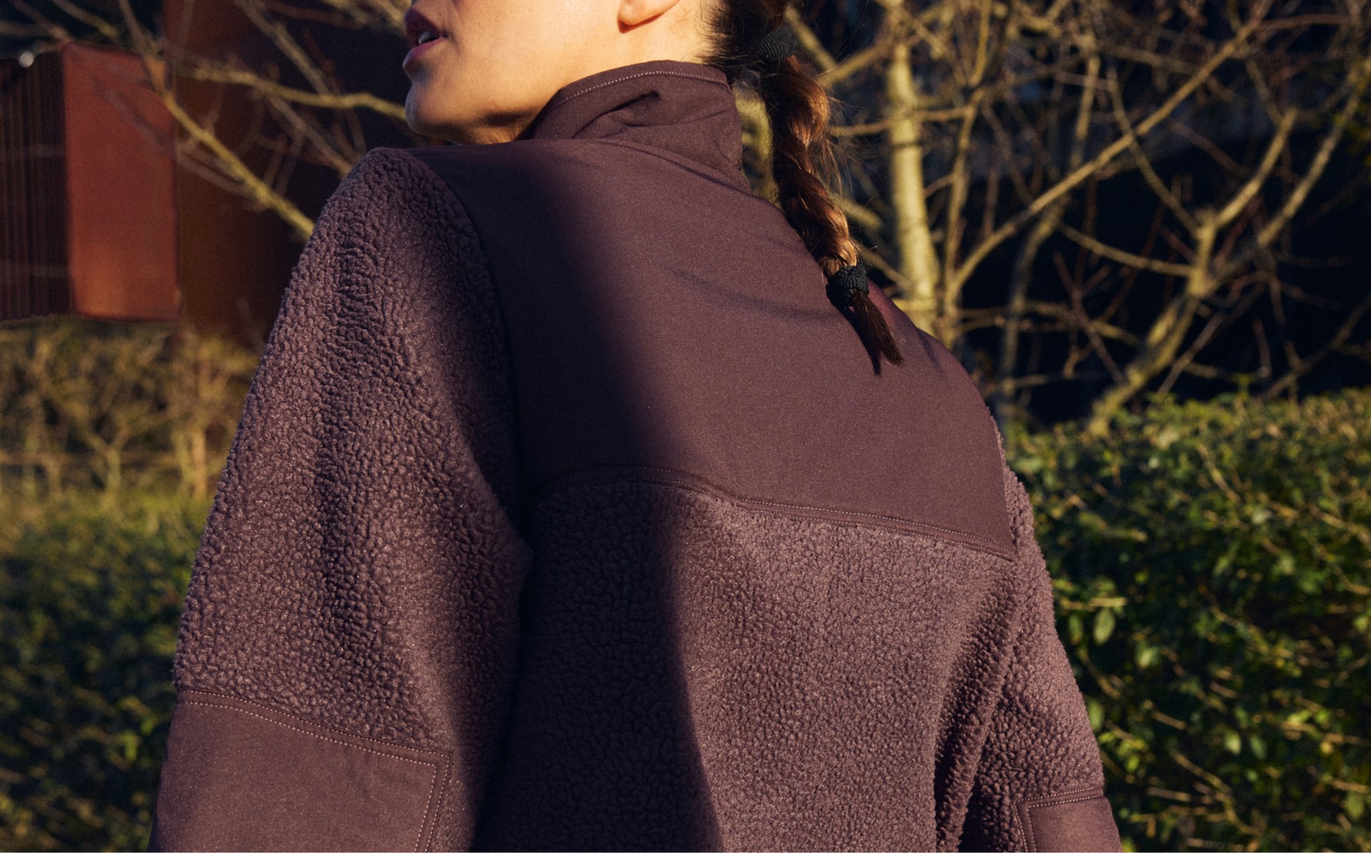 Woman with a braided ponytail holding a yoga mat, wearing a brown fleece jacket, walking outdoors on a sunny day.