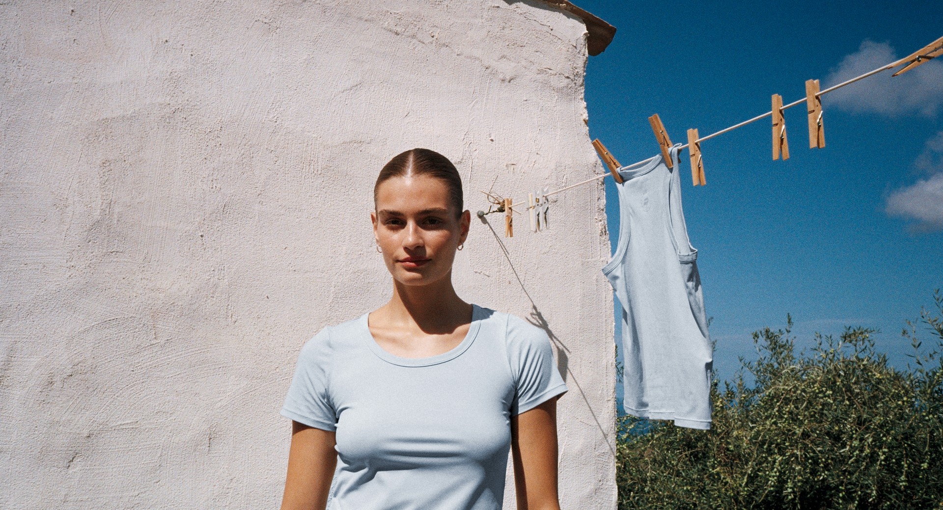 A woman hangs a light blue top on a clothesline outdoors, against a clear blue sky and a rustic white wall.