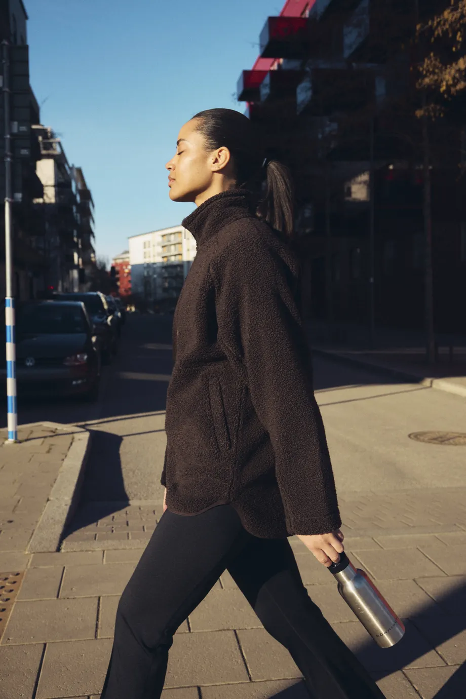 Woman in black fleece walking on a sunny city street, holding a metal water bottle, with buildings and blue sky in the background.