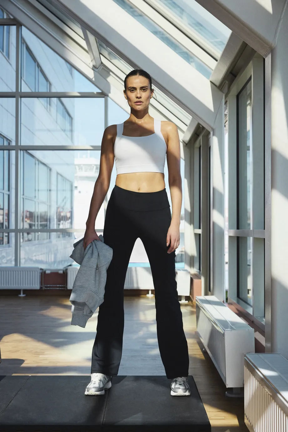 Woman in workout attire stands confidently in a sunlit gym, holding a gray jacket, with large windows in the background.