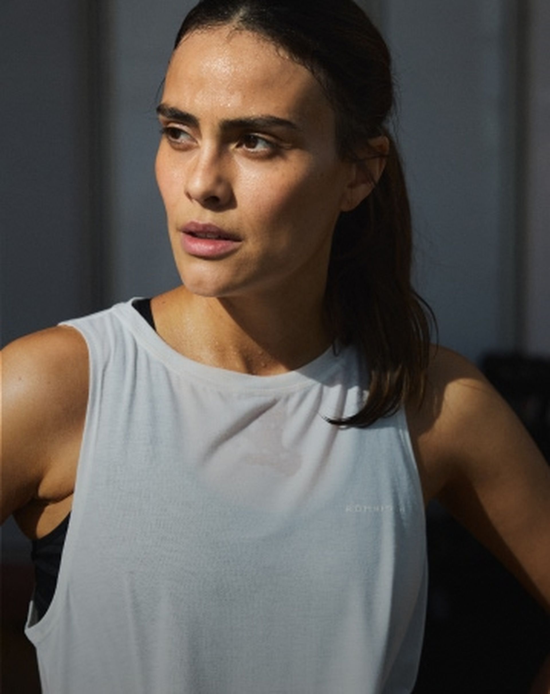 A woman in a sleeveless white top, with her hair tied back, appears focused and slightly sweaty, suggesting she is exercising.
