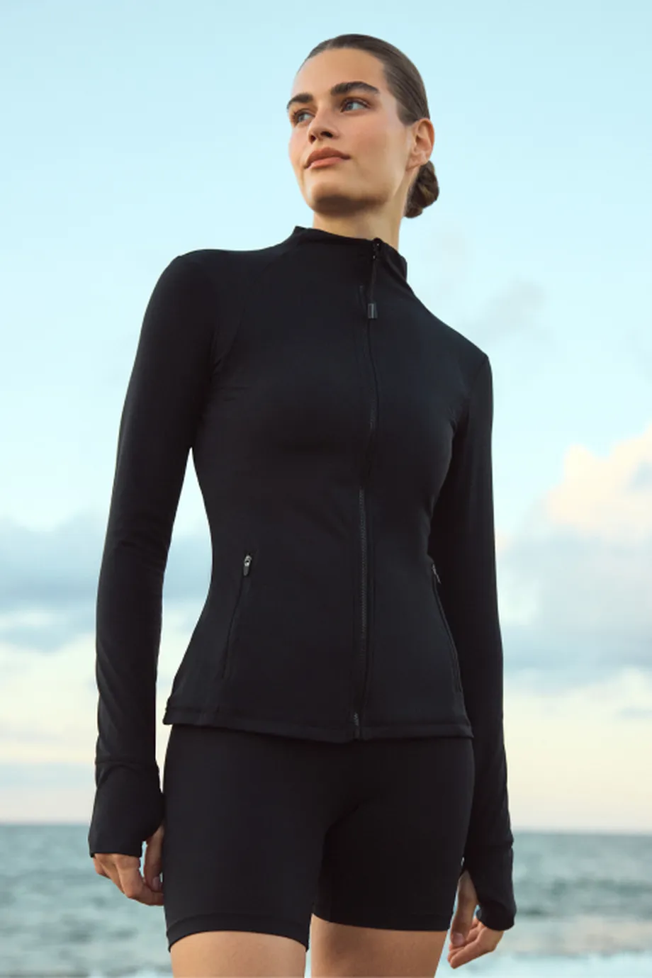 Woman in a black zip-up jacket and bike shorts standing on a beach, looking off to the side under a pale sky.