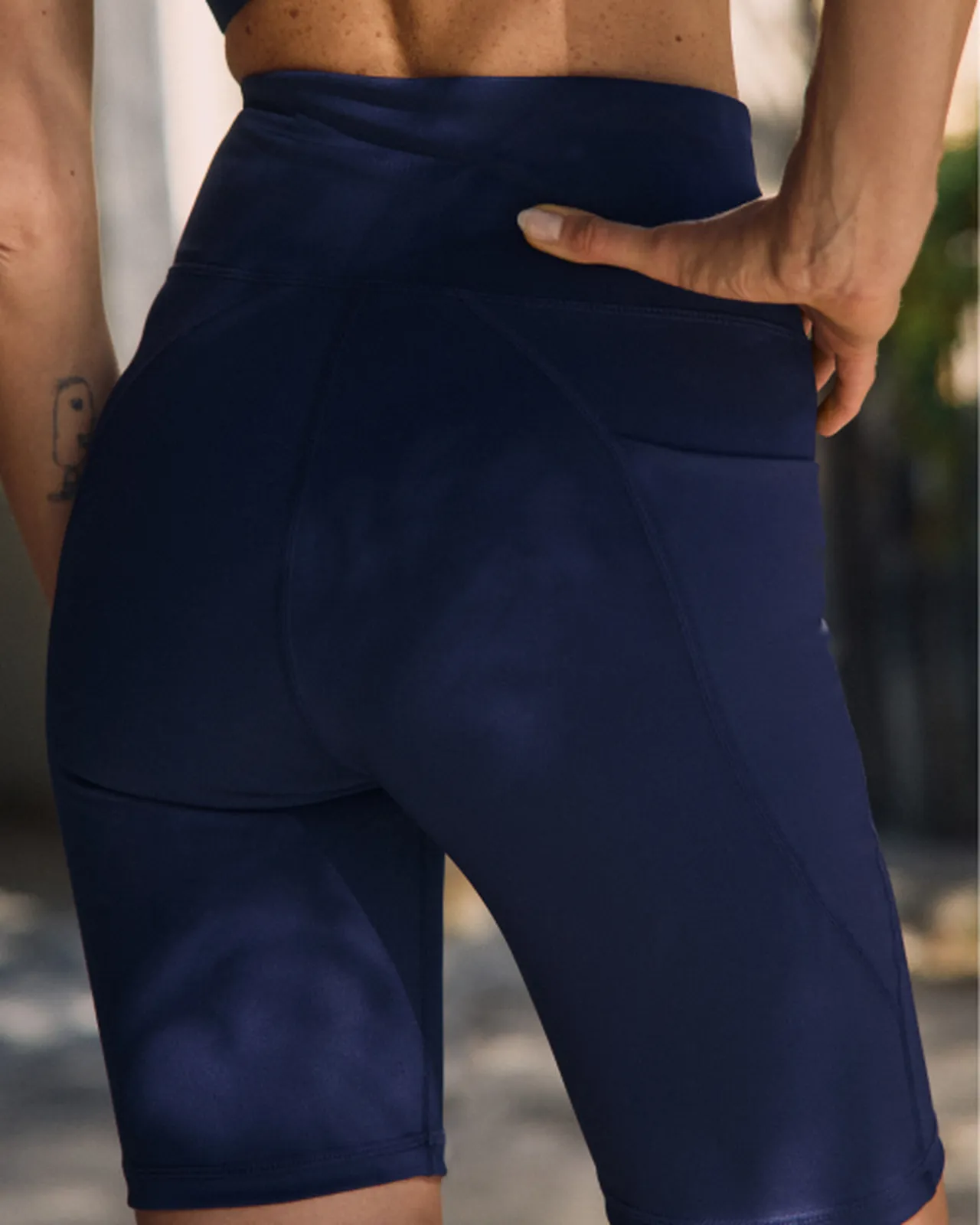 Person in navy athletic shorts standing outdoors, hand resting on back. Sunlight casts soft shadows on their skin.