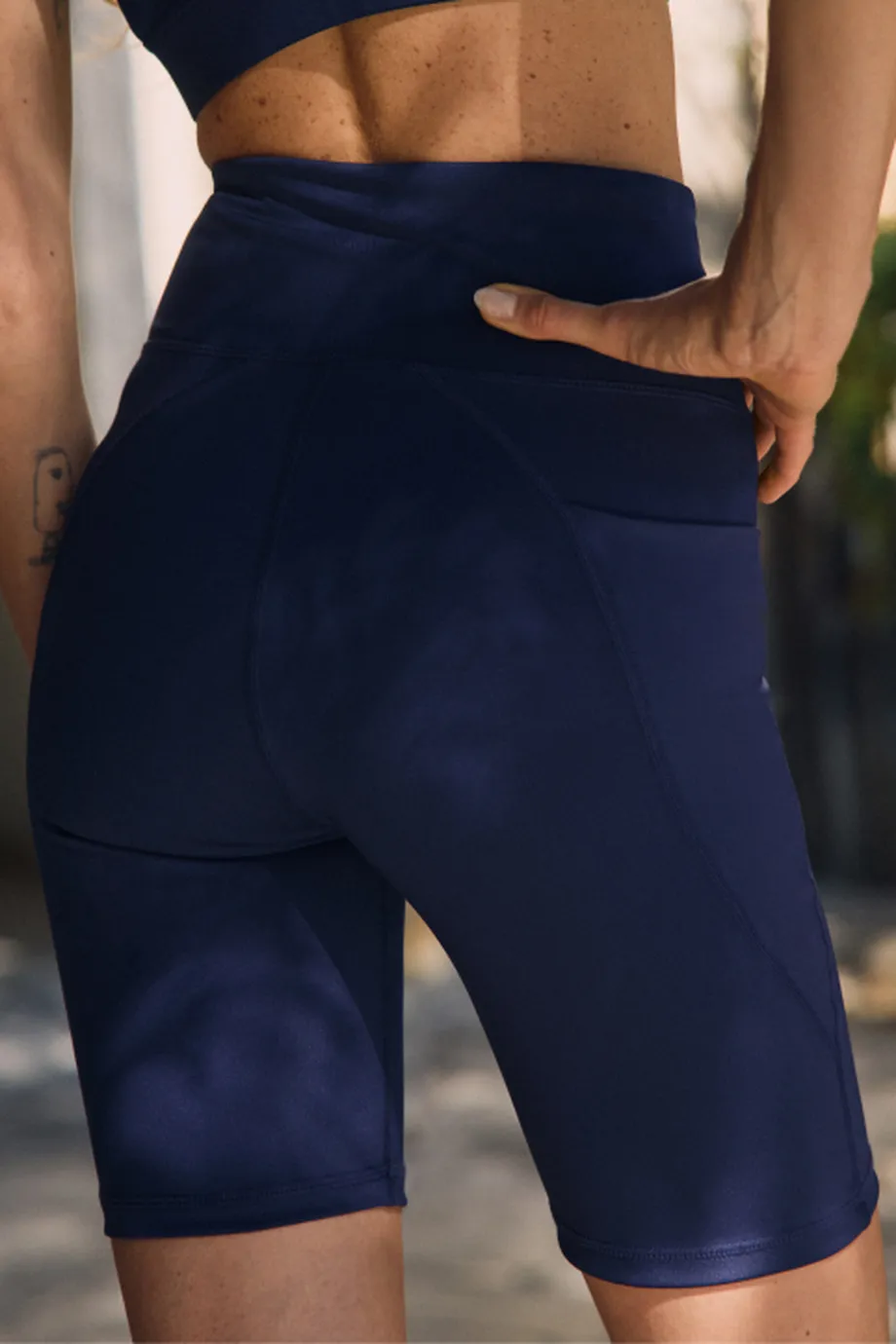 Person in navy athletic shorts standing outdoors, hand resting on back. Sunlight casts soft shadows on their skin.