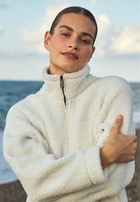 Woman in a white sherpa zip-up jacket, arms crossed and hugging herself, smiling slightly on a beach with ocean and cloudy sky behind.