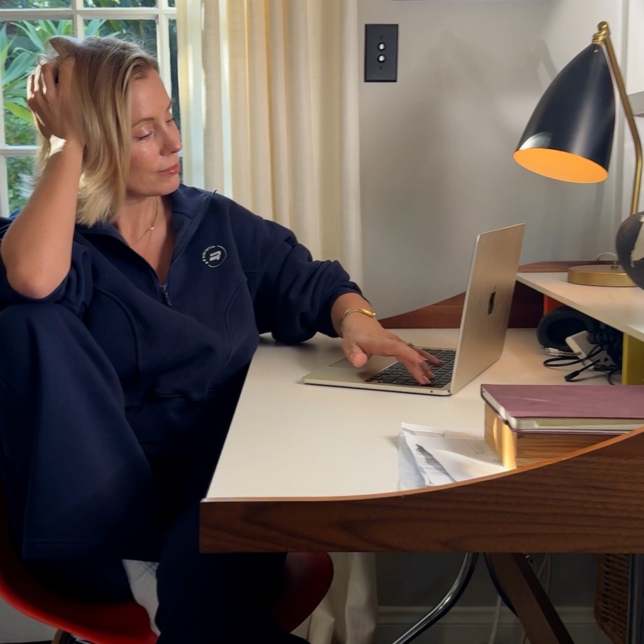 Woman in a navy tracksuit sits at a desk with a laptop, hand in hair, lit by a desk lamp, near a window with beige curtains.