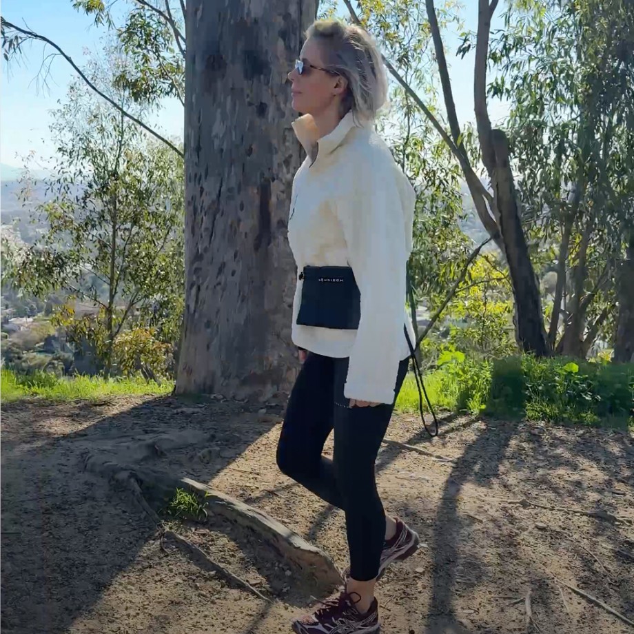 Woman in athletic wear walking on a shaded forest trail, sunlight filtering through tall trees in the background.