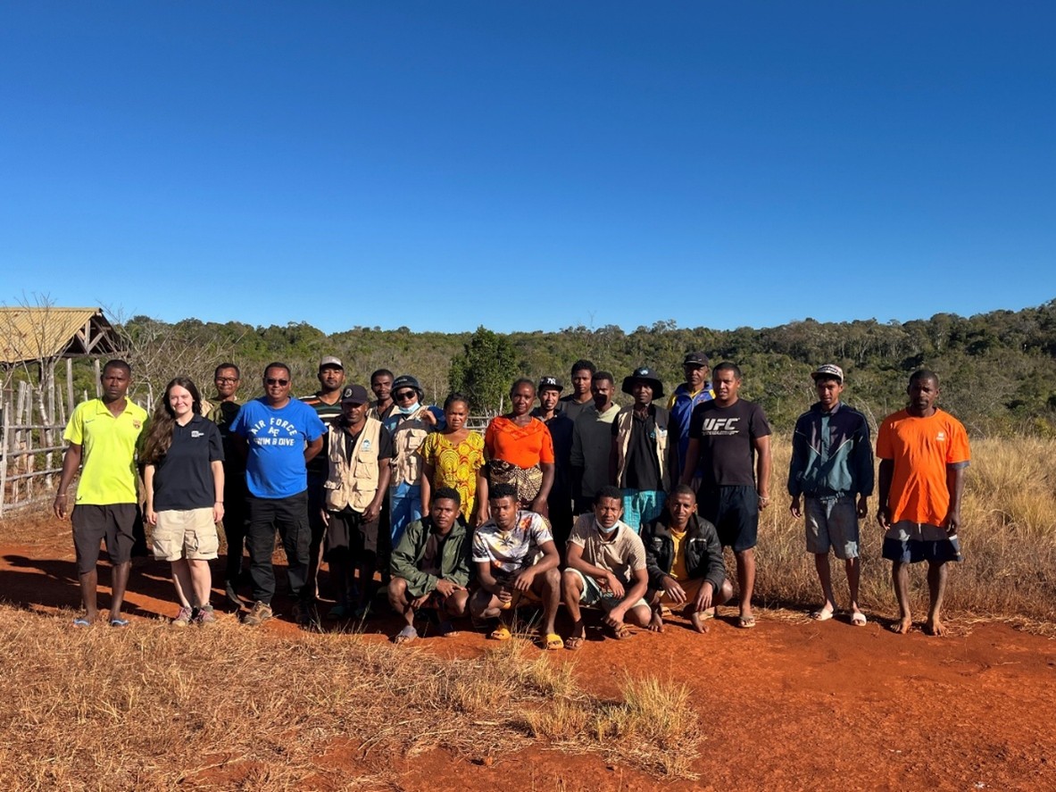 A group of people standing in the African Savannah with a hut behind and a bright blue sky.