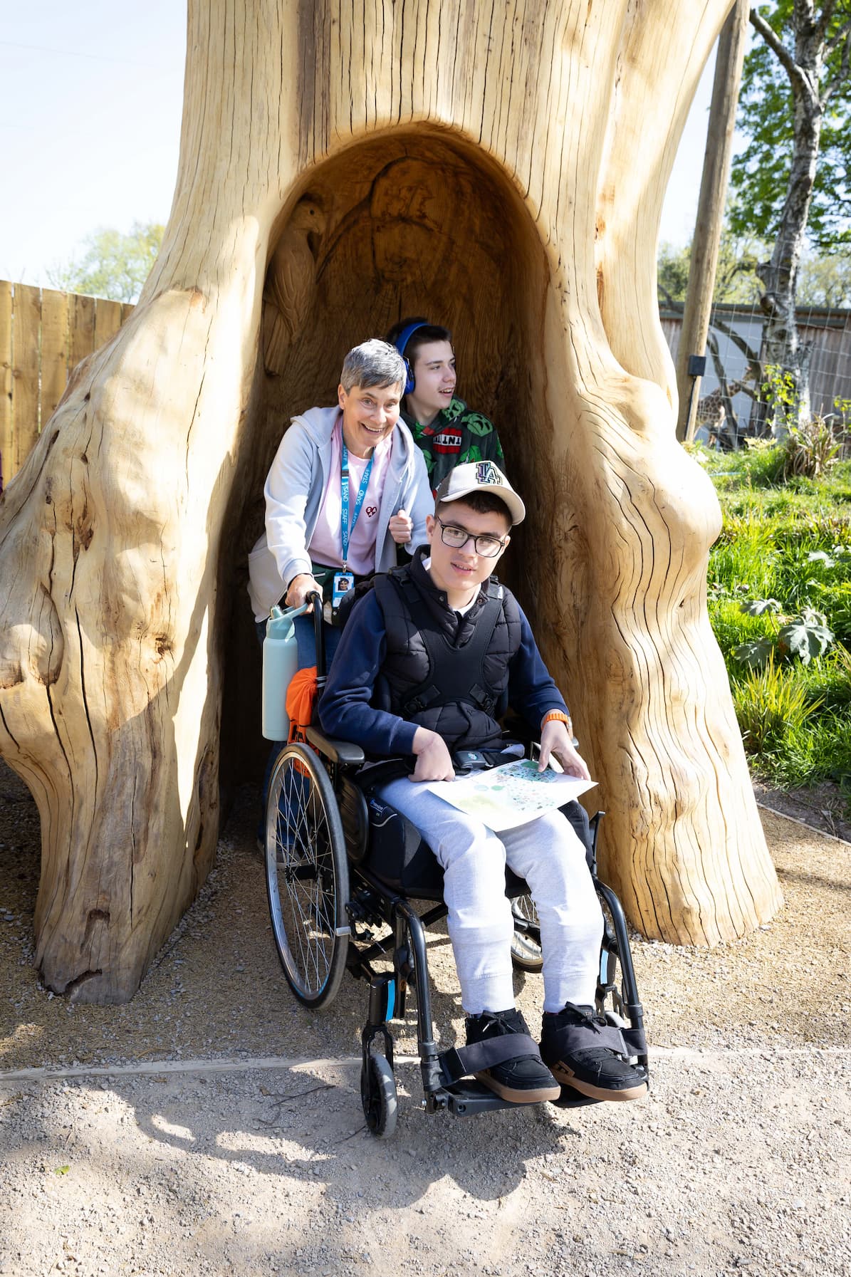 A young person in a wheelchair holding a map, a smiling adult, and another child pose inside a large wooden sculpture outdoors.