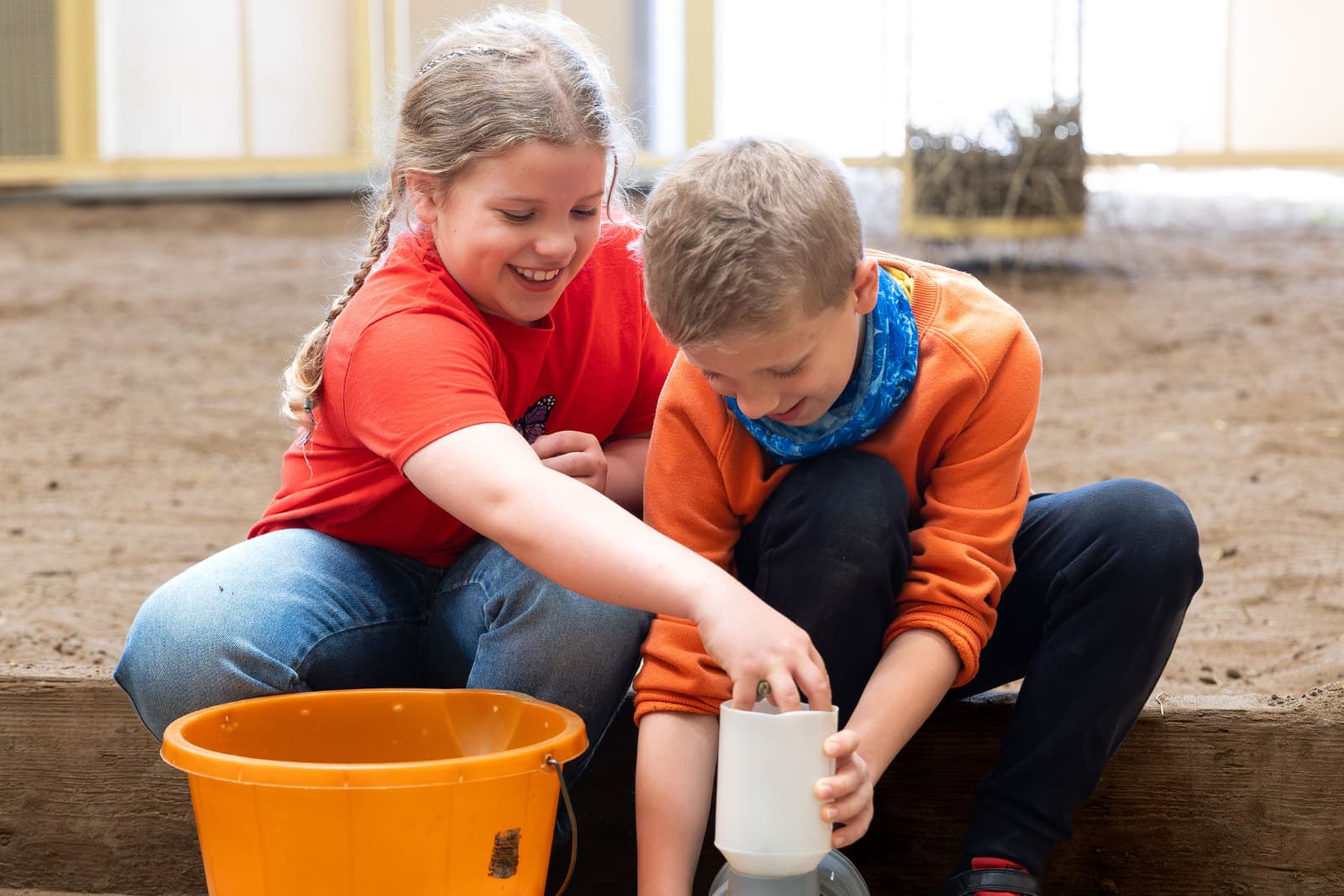 Giraffe Experience Two smiling children sitting on a wooden ledge, playing with a white container and an orange bucket in a sandy indoor area.