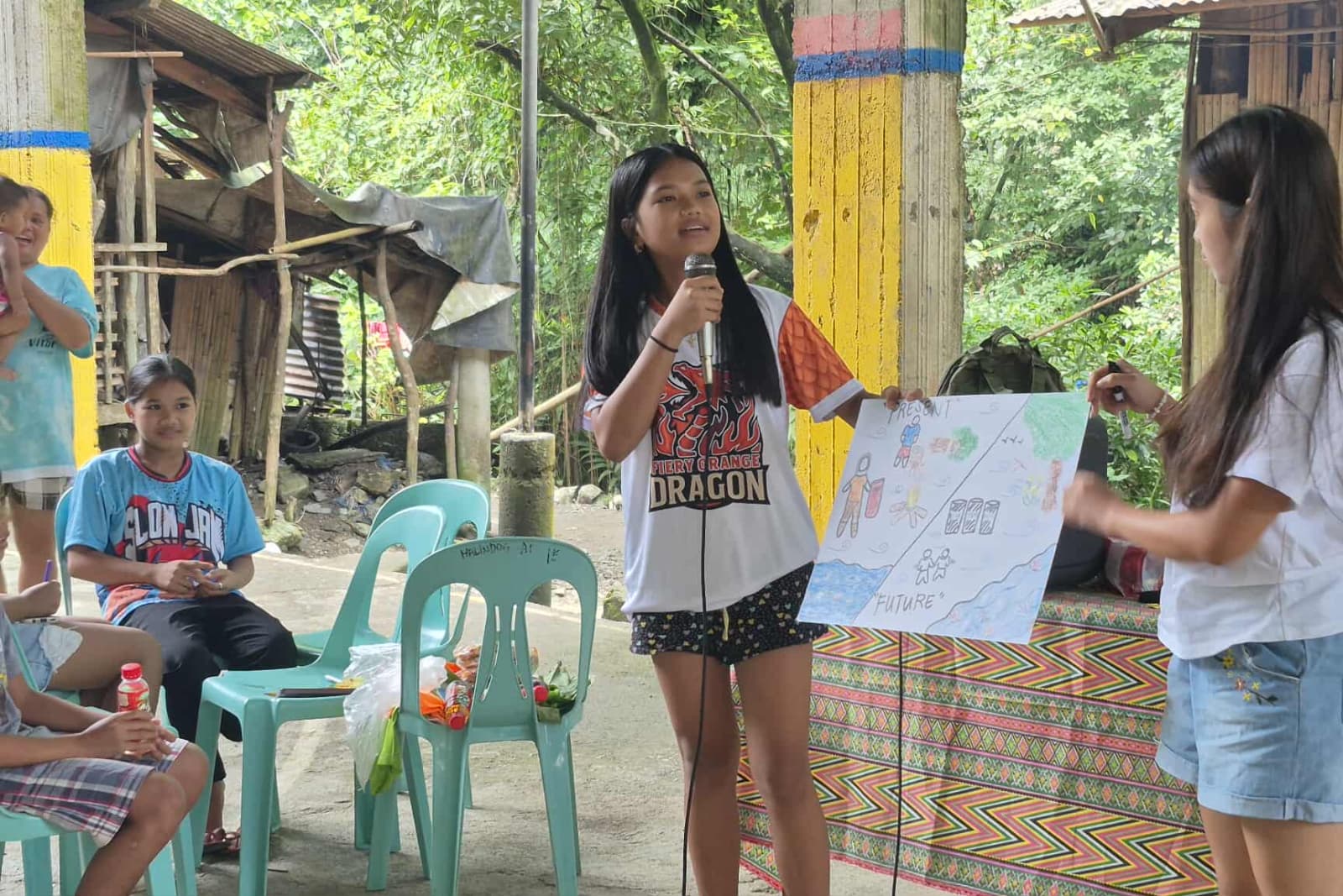 Philippines Girl presenting environmental poster outdoors, using a microphone while others sit and watch, among chairs and colorful surroundings.