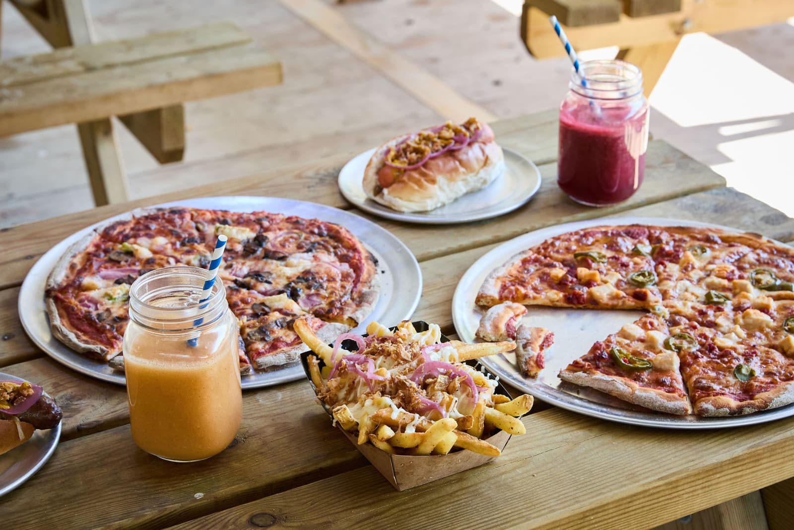 Base Camp Pizzeria Pizzas, a hot dog, loaded fries, and smoothies on a wooden picnic table outdoors.