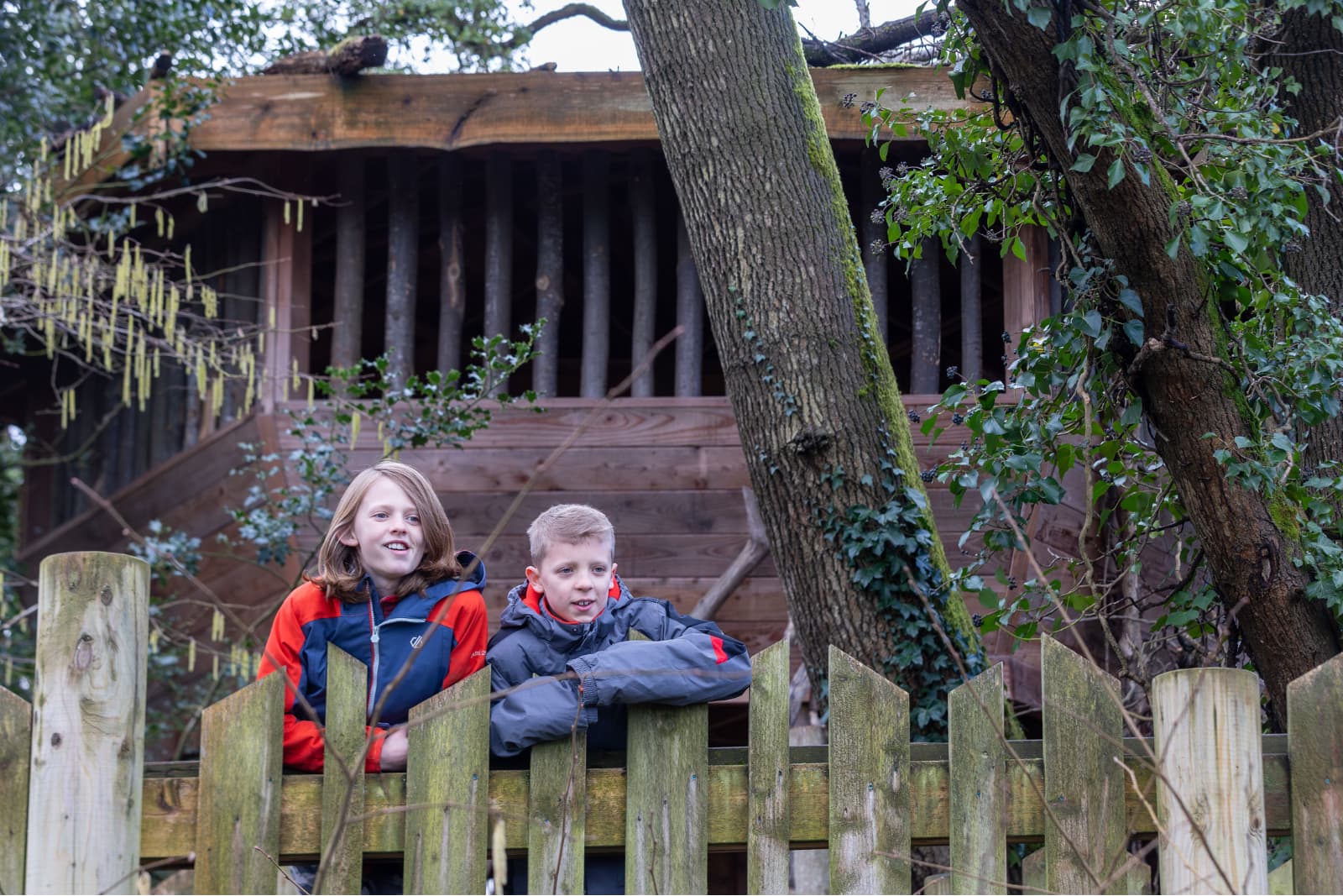 Kids in Bear Wood Two children in jackets leaning on a mossy wooden fence in front of a rustic treehouse surrounded by trees and ivy.