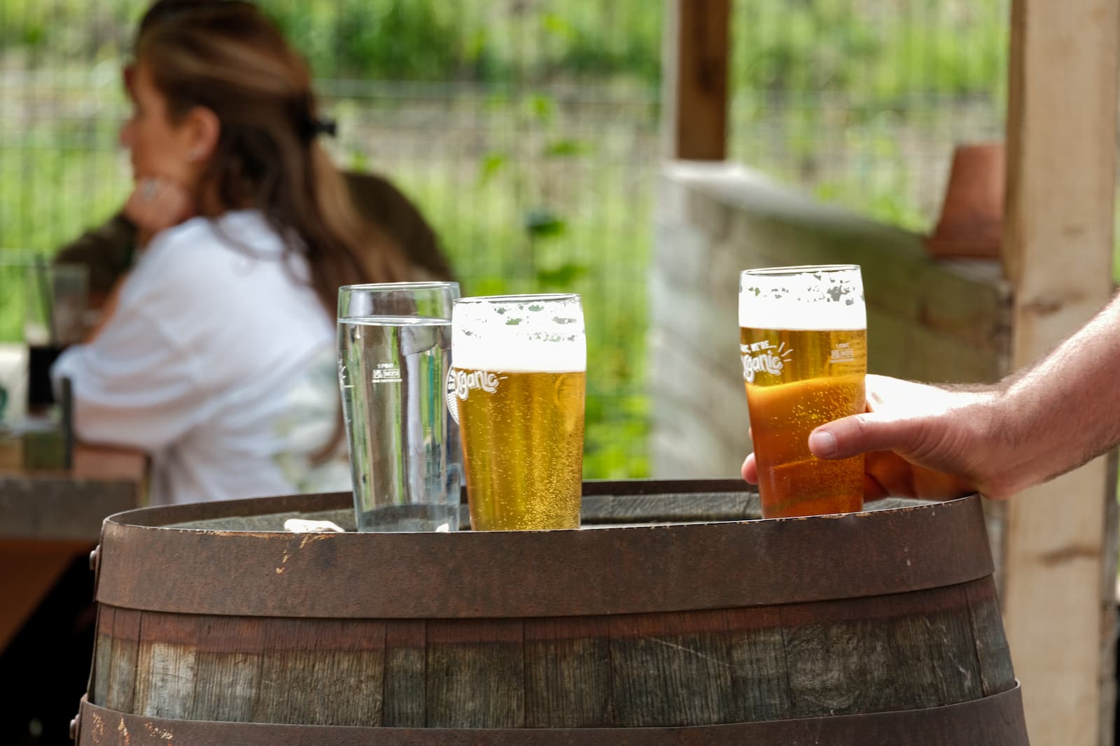 A close up of pints of beer resting on a wooden barrel, with a hand stretching out to hold one