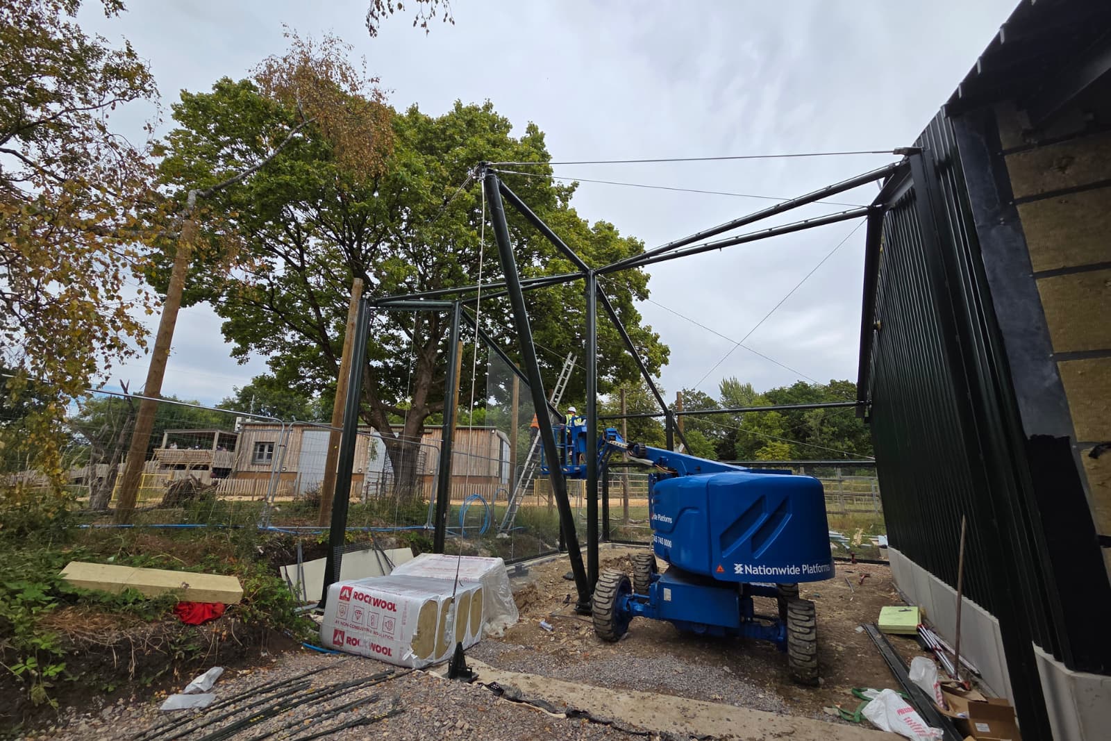 Construction site with a blue lift, metal framework, and building materials. Trees and a partly cloudy sky are in the background.