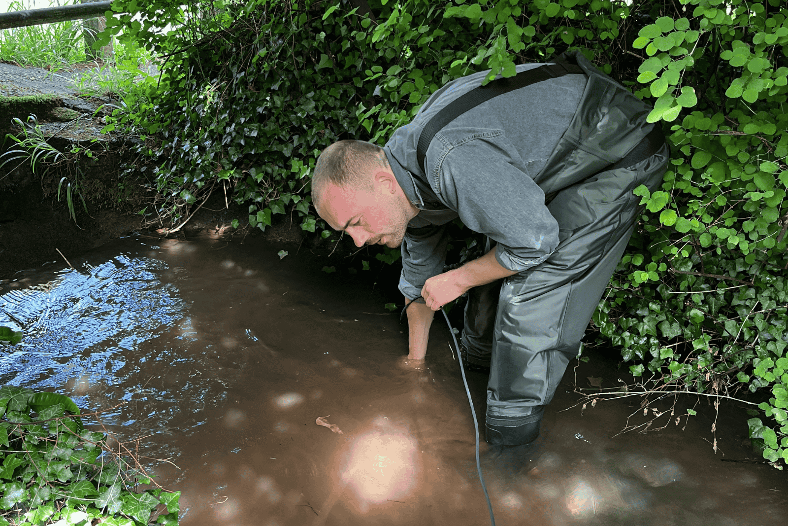 An individual wearing waders crouches in a river