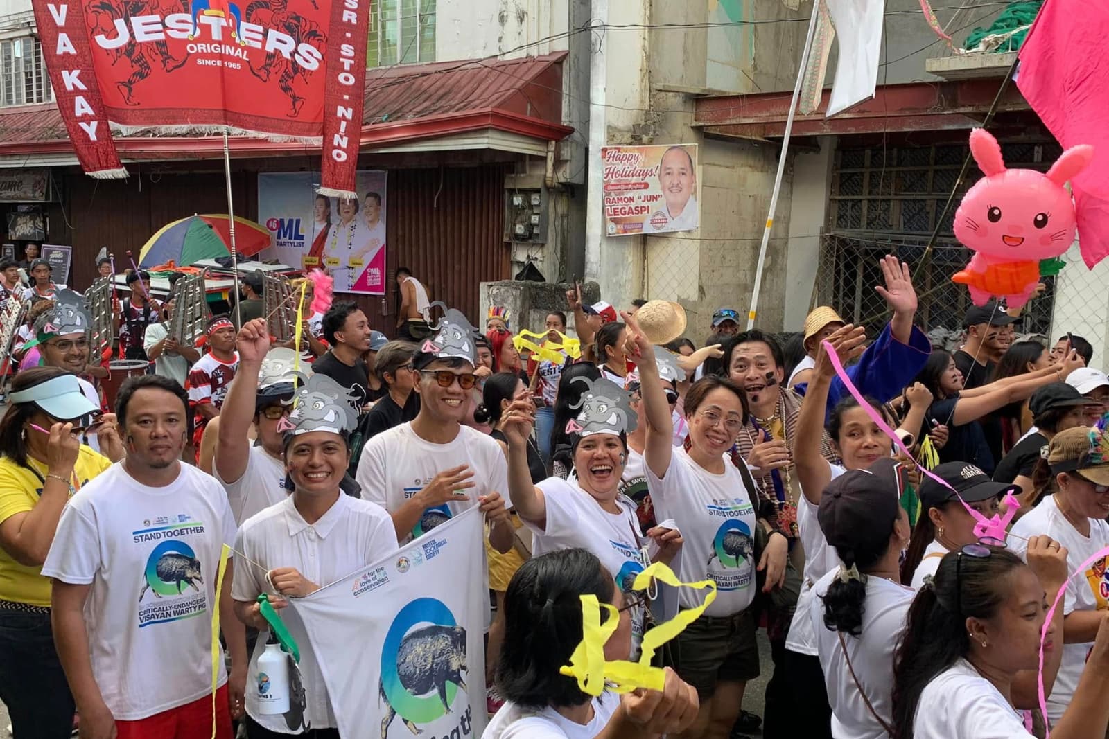 A lively group of people in white shirts and hats celebrate outdoors with banners, balloons, and colorful decorations.