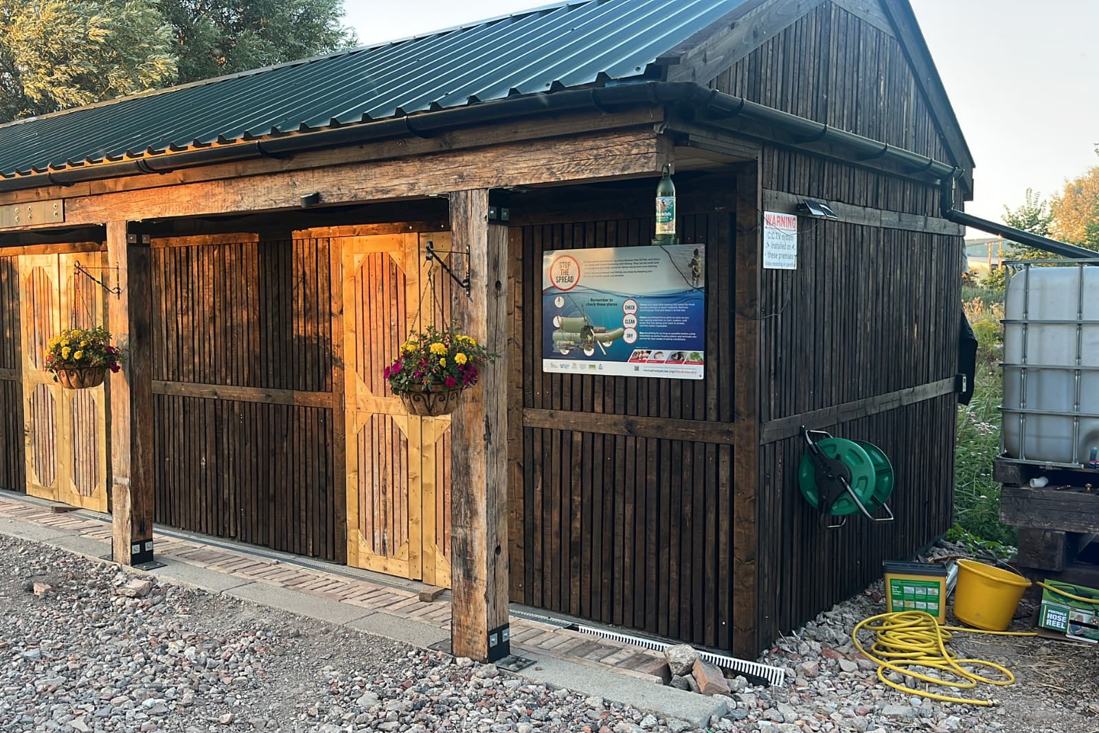 AQUA scheme Wooden shed with two hanging flower baskets, a poster on the side, a green hose reel, and various outdoor items on the ground.