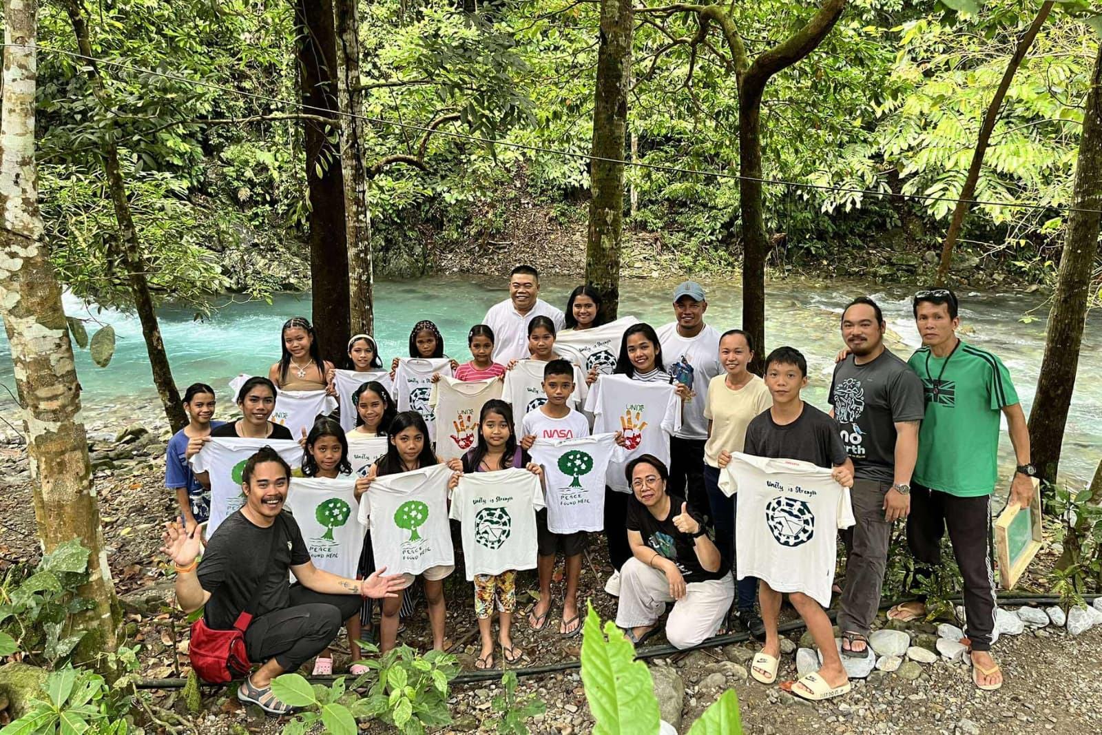 Philippines Group of people holding T-shirts with environmental designs, posing in a lush forest by a clear blue river.
