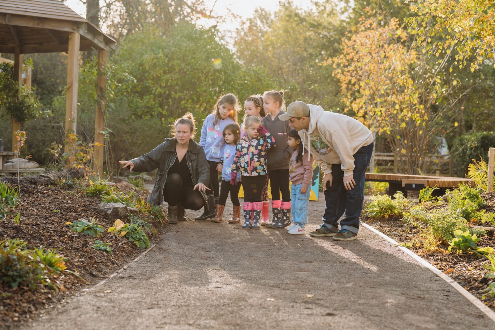 Rainforest garden A group of children and two adults explore a garden path, with one adult pointing at something on the ground. Trees and a gazebo are in the background.