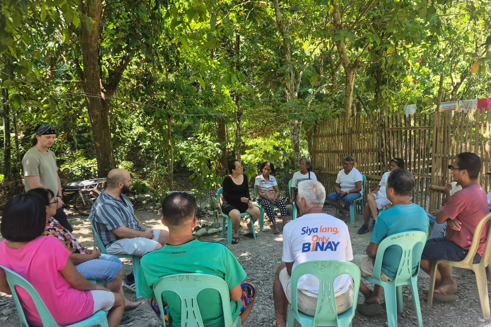 Philippines A group of people sit in a circle of plastic chairs in an outdoor setting, surrounded by trees and bamboo fencing.