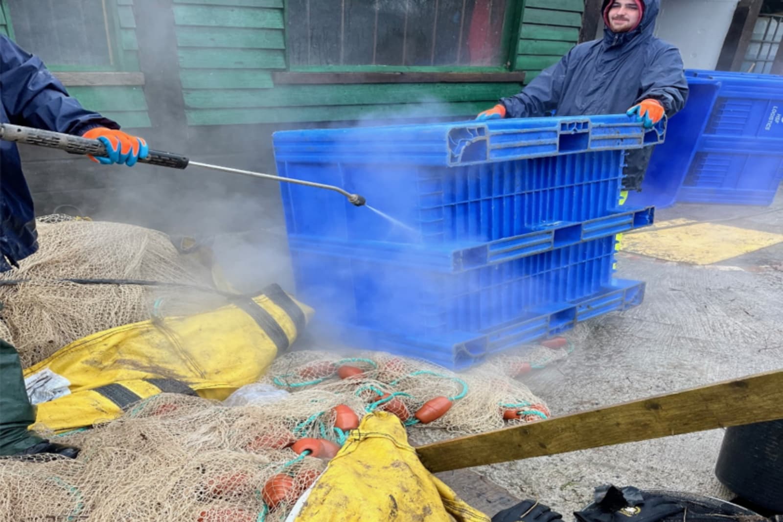 AQUA Workers pressure-wash blue crates near fishing nets and gear outside a green building.