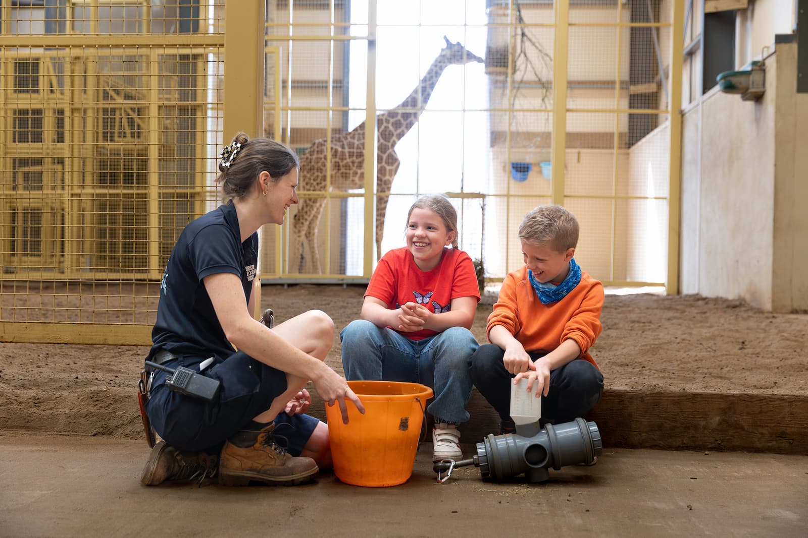 A zookeeper crouches in an animal habitat with two children, with a giraffe in the background