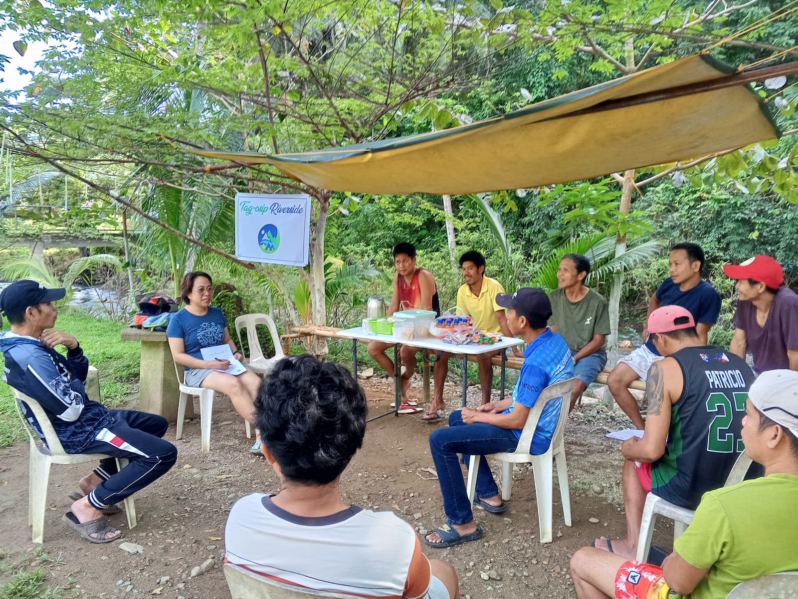 A group of people sit in a semi-circle outdoors under a canopy, engaged in a discussion beside a 