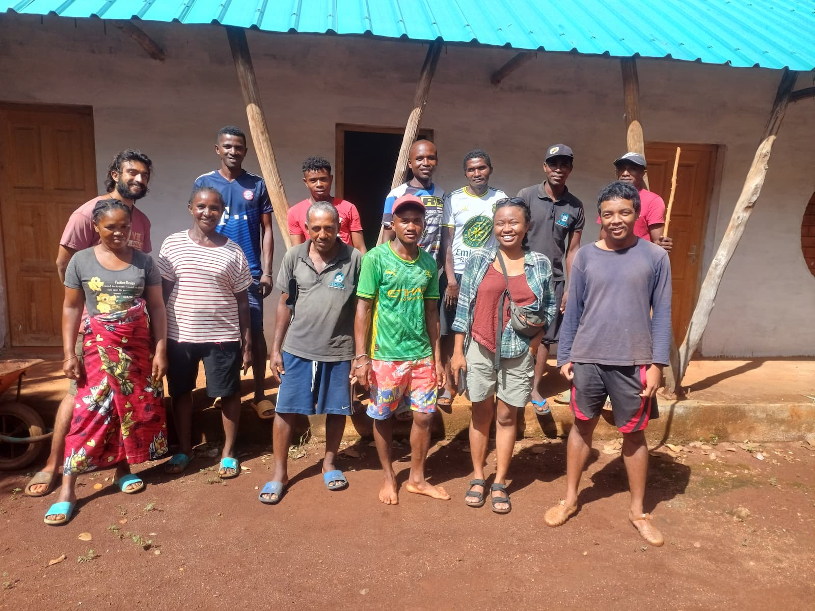 A group of thirteen people standing in front of a building with a blue roof, smiling at the camera. The ground is reddish-brown soil.