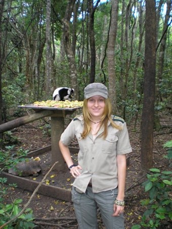 A woman standing in front of a ruffed lemur on a eating station in the middle of a jungle