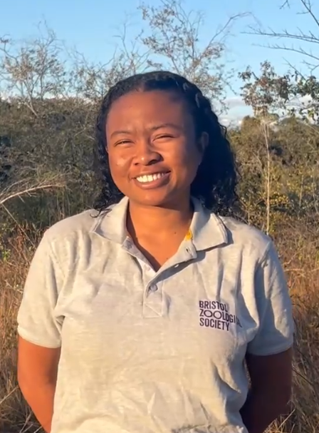 A lady standing smiling to camera with a Bristol Zoological Society polo shirt on and foilage in the background