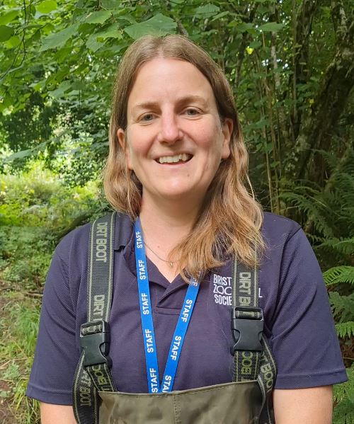 A person with long hair, wearing a Bristol Zoological Society shirt and staff lanyard, stands outdoors surrounded by lush green foliage.