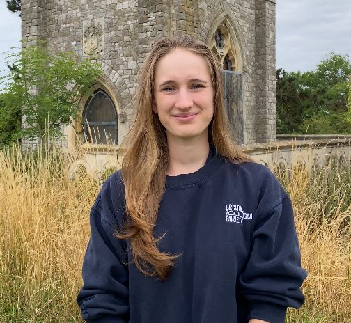 A person with long hair stands in front of a stone building, wearing a navy sweatshirt, surrounded by tall grass.