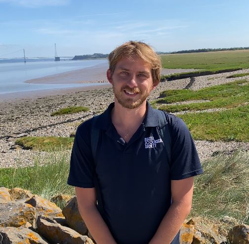 A person with a beard stands smiling near a rocky shoreline, with a grassy landscape and a bridge in the background under a clear blue sky.