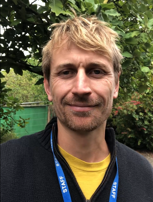A man with blond hair and a beard smiles at the camera, wearing a black sweater and a blue lanyard, standing outdoors near greenery.