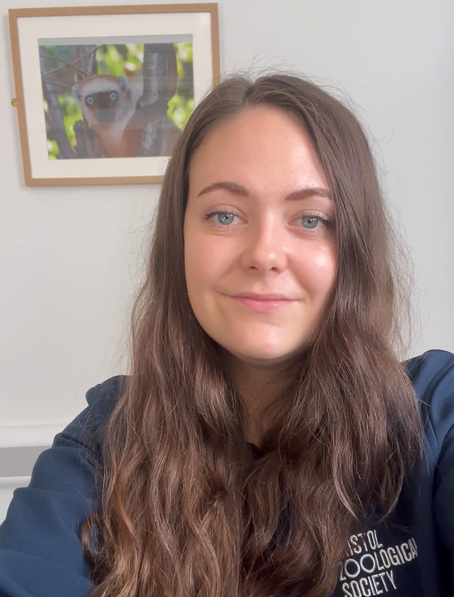 A woman with long brown hair smiles at the camera. Behind her is a framed picture of a lemur on the wall.