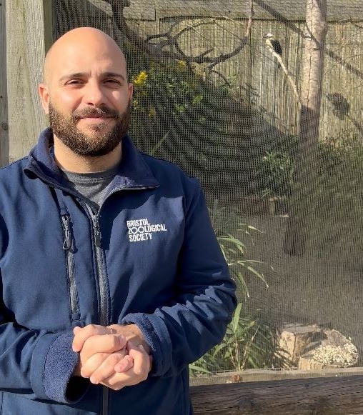 A person standing in front of a bird aviary wearing Bristol Zoological Society uniform