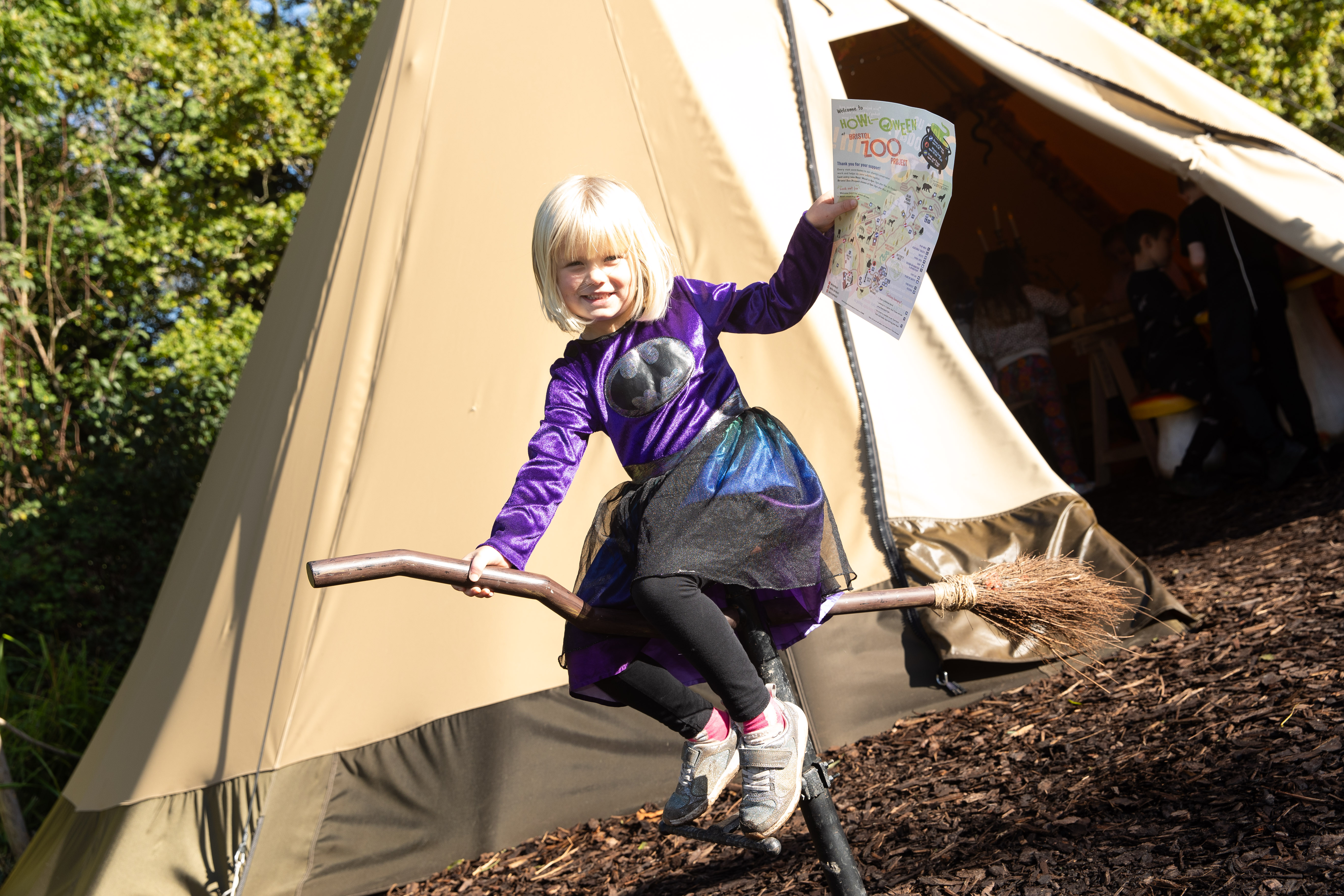 Child in a purple witch costume sits on a broomstick in front of a teepee, holding a map and smiling.