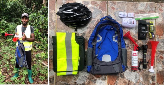 A person in a safety vest holds an air horn. Nearby, a helmet, backpack, safety vest, and various tools are laid out on a tiled surface.