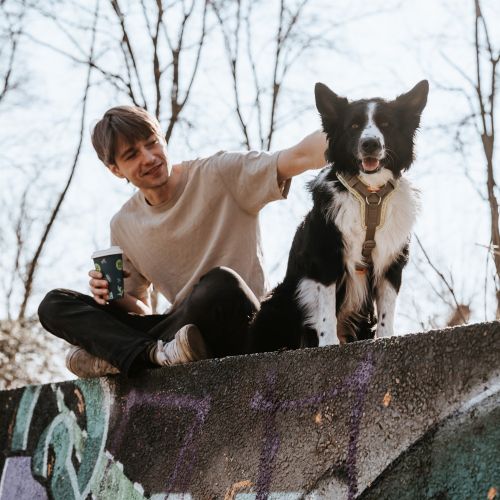 Ein Mann sitzt auf einer mit Graffiti übersäten Mauer, lächelt und streichelt einen schwarz-weißen Hund. Im Hintergrund sind Bäume mit kahlen Ästen zu sehen.