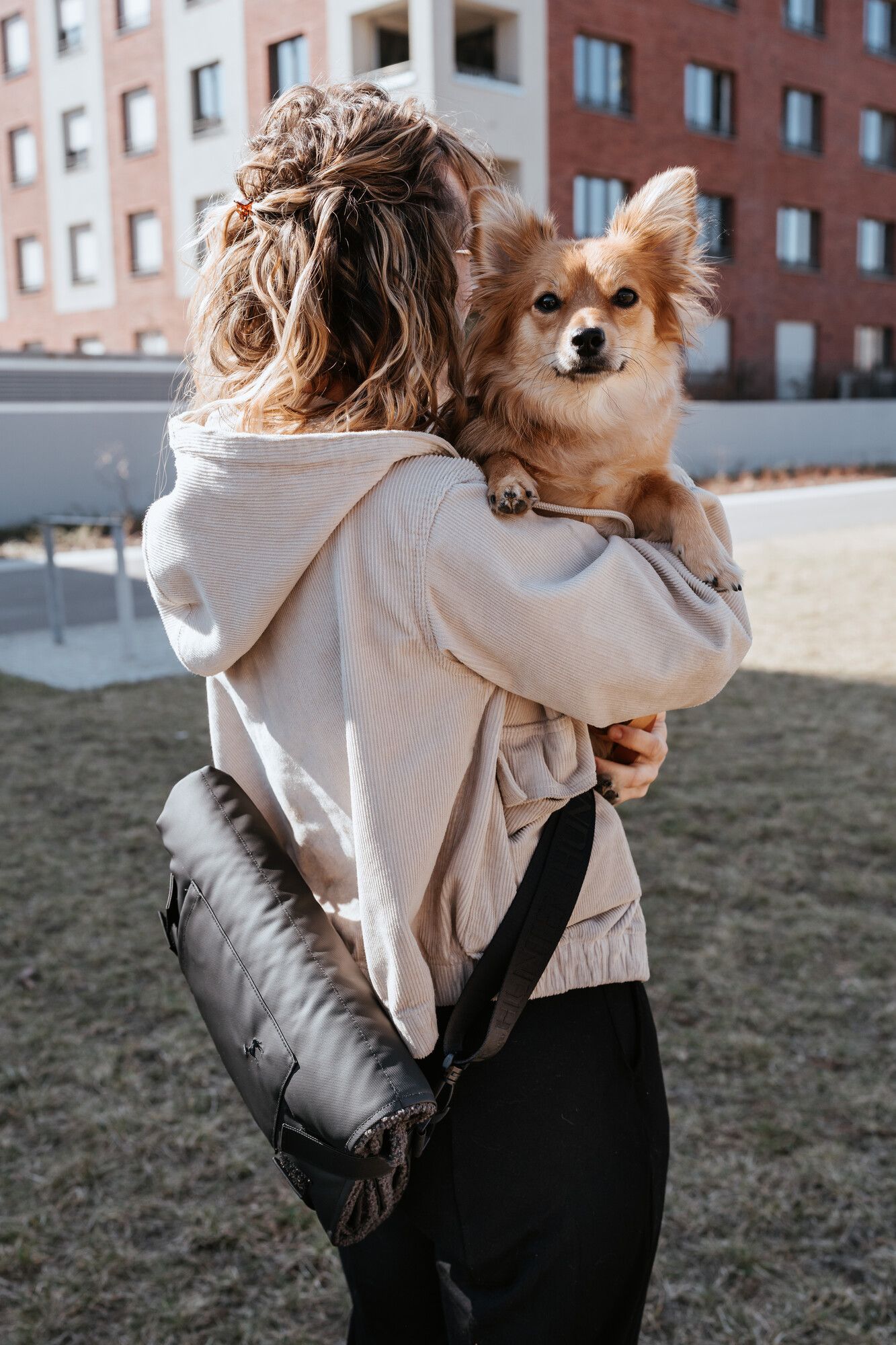 Eine Person mit gewelltem Haar hält draußen einen kleinen braunen Hund im Arm. Sie trägt einen beigen Kapuzenpulli und hat eine schwarze Tasche dabei. Im Hintergrund sind Wohnhäuser zu sehen.