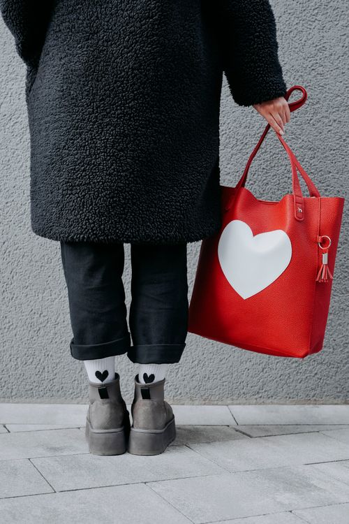Person in schwarzem Mantel und schwarzer Hose, die eine rote Tragetasche mit einem großen weißen Herz hält. Sie trägt Socken mit schwarzen Herzmotiven und graue Schuhe.