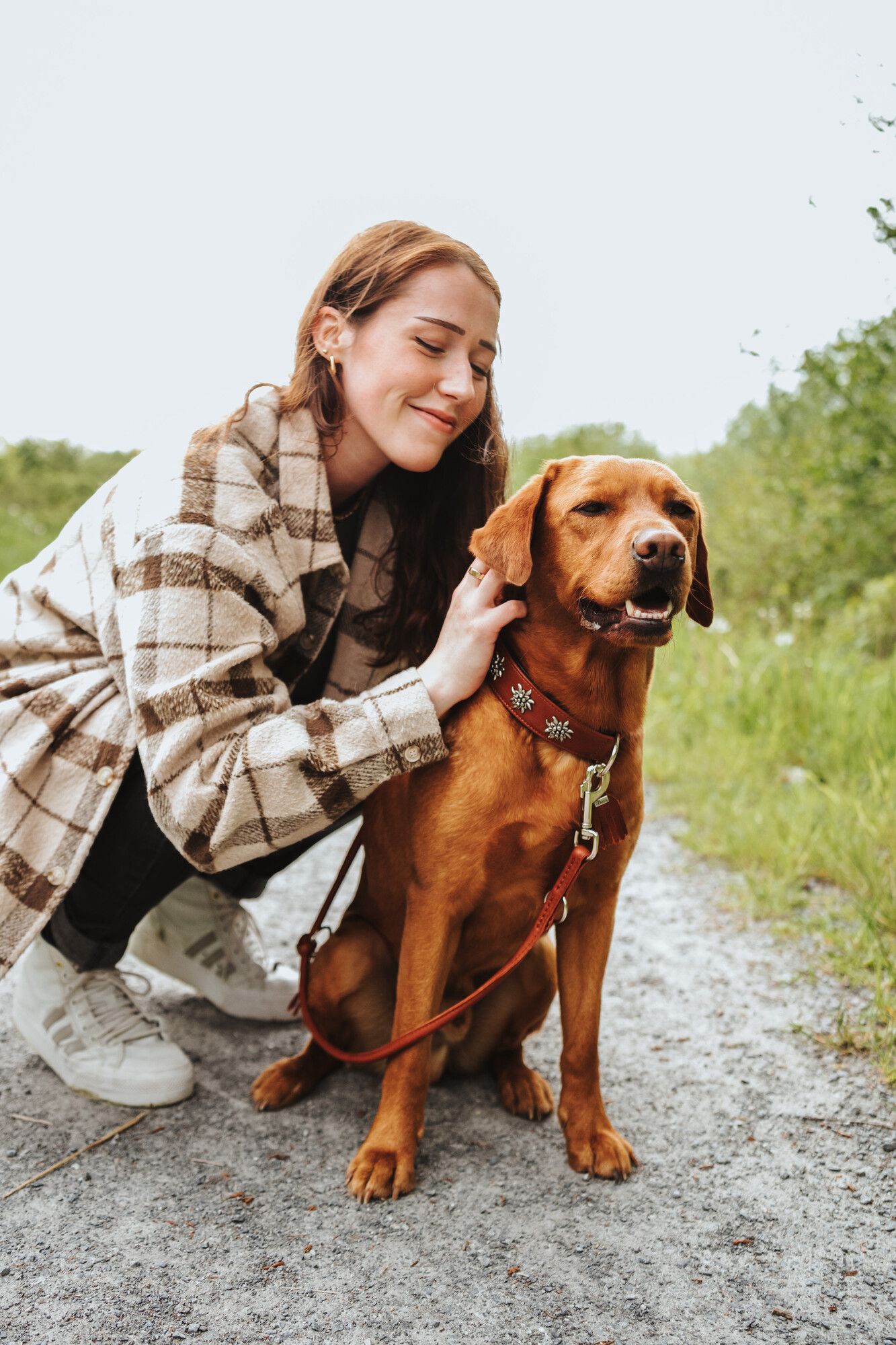 Eine Frau in einem karierten Mantel kniet neben einem fröhlichen braunen Hund an der Leine, draußen auf einem Kiesweg mit Grünpflanzen im Hintergrund.