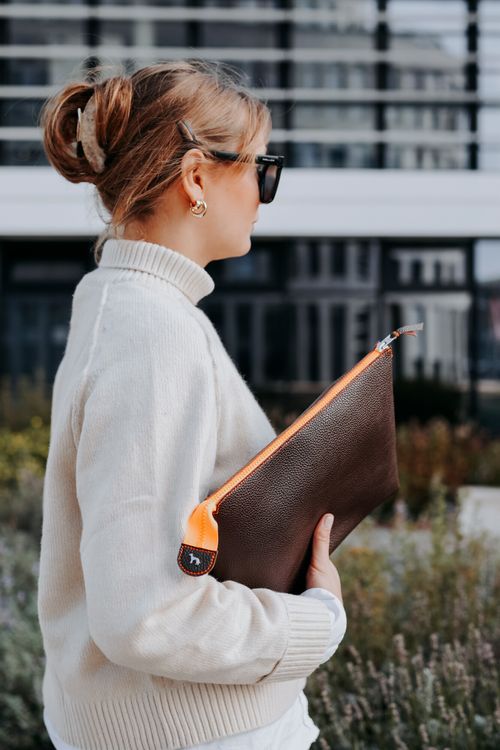 Frau mit Sonnenbrille und beigem Rollkragenpullover hält eine braunen Kulturbeutel mit orangefarbenem Reißverschluss in der Hand und steht im Freien in der Nähe eines modernen Gebäudes.