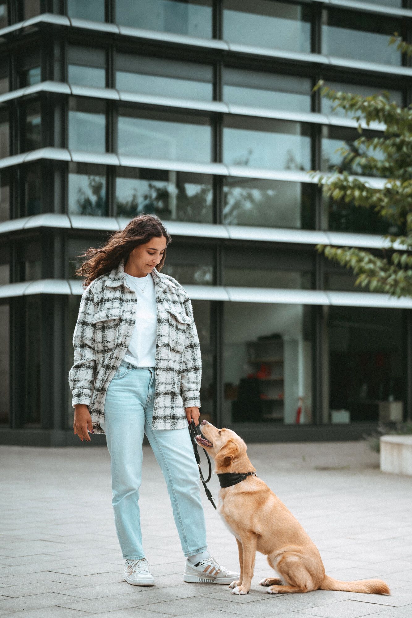 Eine Frau in einer karierten Jacke steht mit einem sitzenden Hund an der Leine vor einem modernen Glasgebäude.