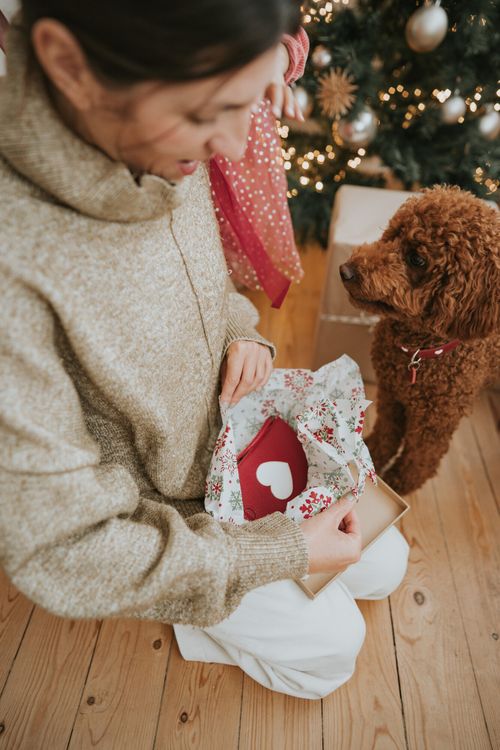 Eine Frau öffnet ein mit einem Herz verziertes Geschenk, während sie neben einem Weihnachtsbaum sitzt und von einem braunen Hund beobachtet wird.