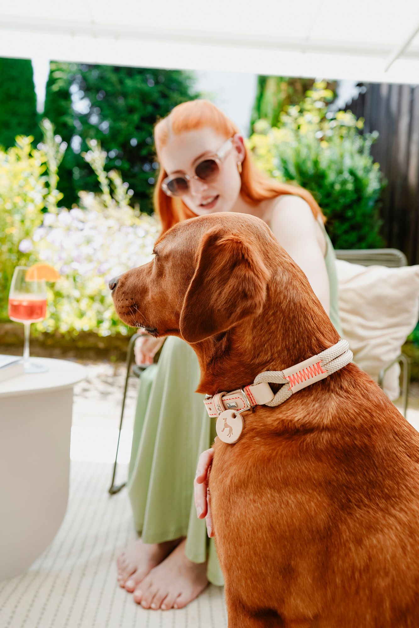 Eine Frau mit roten Haaren und Sonnenbrille sitzt im Freien und streichelt einen braunen Hund. Auf dem Tisch neben ihnen steht ein Getränk.