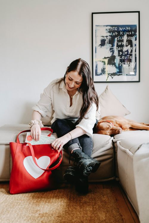 Eine Frau sitzt auf dem Sofa mit einer großen roten Tasche, auf der ein weißes Herz abgebildet ist. Ein Hund schläft neben ihr, während sie die Tasche mit einem fröhlichen Gesichtsausdruck betrachtet.